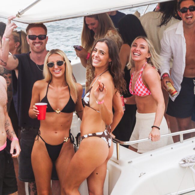 A group of people enjoy a party on a boat, smiling and posing for the camera. Two women in bikinis stand at the front, holding drinks, while others chat and laugh in the background near the water.