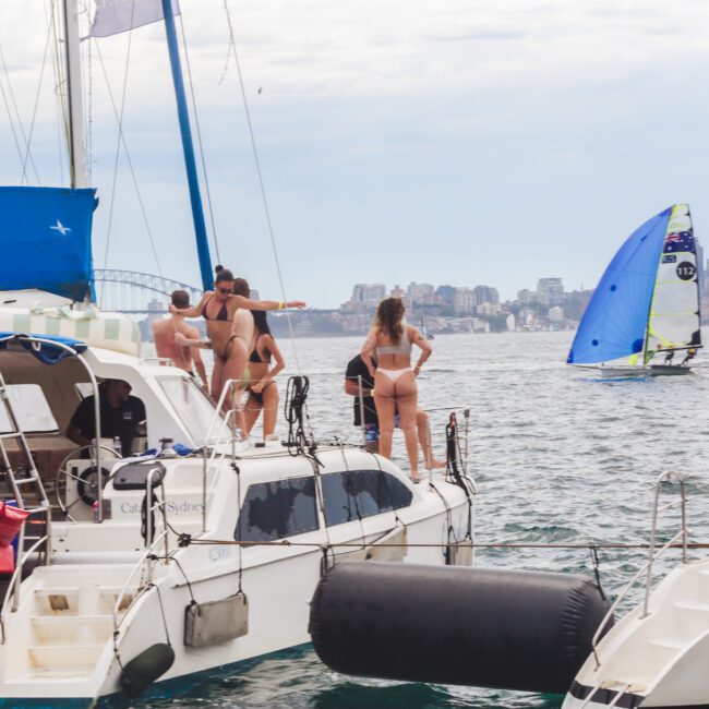 A group of people in swimwear stand on the deck of a catamaran, watching a sailboat race in the water. Other boats float nearby, with a city skyline and cloudy sky in the background.