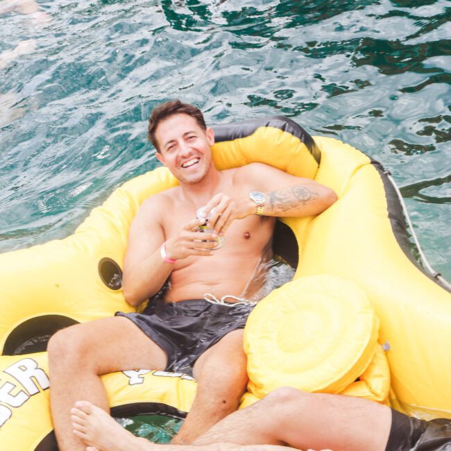 A smiling man in swim trunks lounges on a yellow inflatable raft in the water, holding a drink. Another person's legs are visible on the raft nearby. The water around them is clear and blue-green.