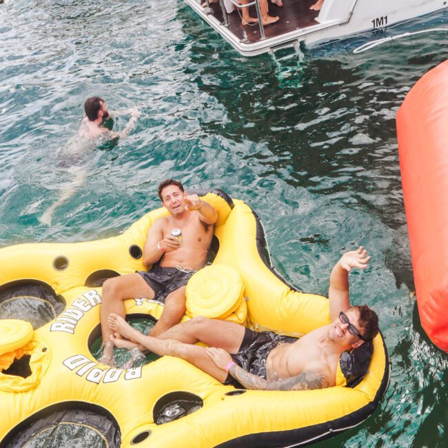 Two men in swim trunks relax and smile on a large yellow inflatable float in the water near a boat party, with people on a boat and swimmers enjoying the festive atmosphere around them.