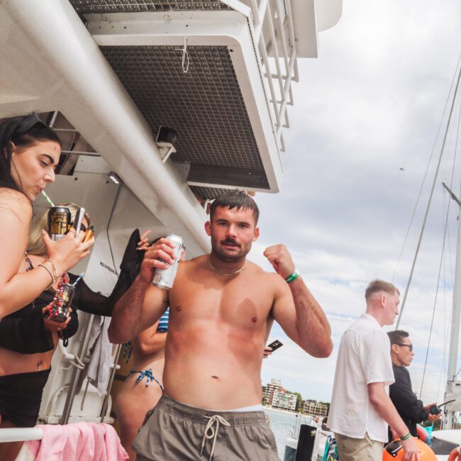 A shirtless man in shorts poses with his fists up, holding a drink, on a boat with other people around him. Some are holding drinks and a trophy. The sky is cloudy and a cityscape is visible in the background.