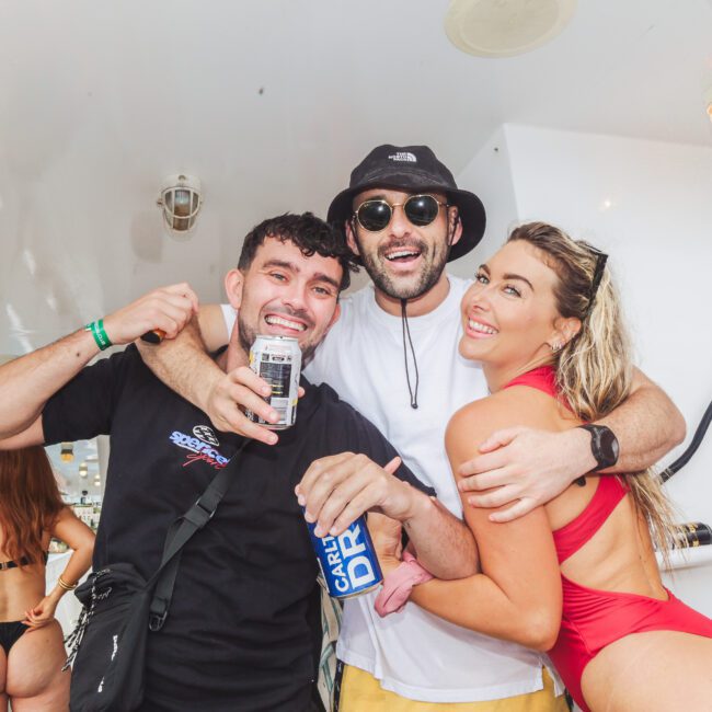Three people smile and pose together on a boat; one man holds a drink, another wears sunglasses and a bucket hat, and a woman in a red swimsuit leans in, with others and water visible in the background.