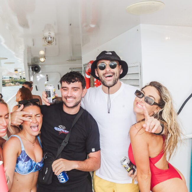 Five young adults in swimsuits and summer clothes smile and pose together on a boat, holding drinks and making playful gestures, with bright daylight and water visible in the background.