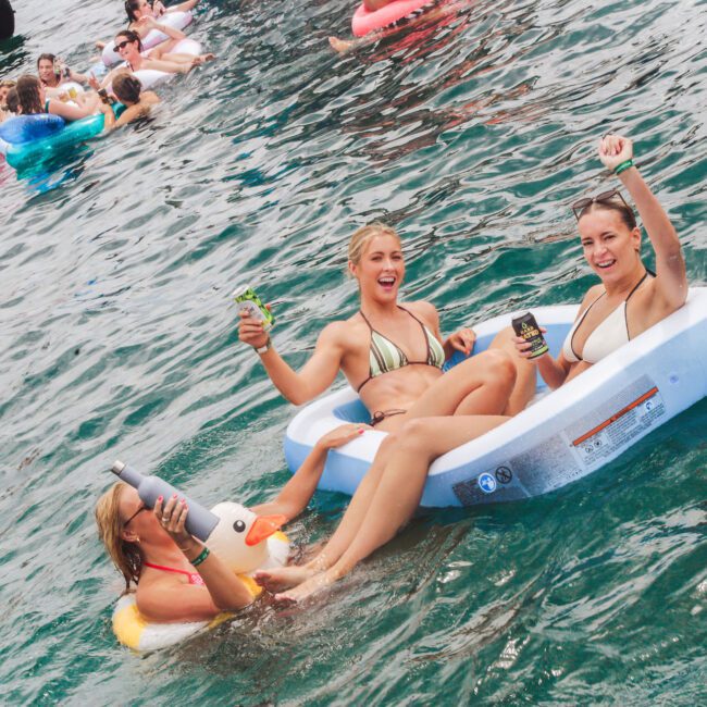 Three women in swimsuits relax on inflatable floats in the water, smiling and holding drinks. Other people on floats are swimming in the background, creating a lively, fun atmosphere.