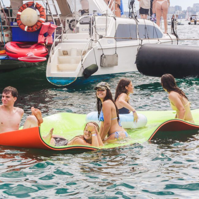 A group of five young adults in swimwear relax on a floating mat in the water near several boats, with a city skyline visible in the background.