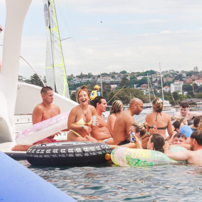 A group of people in swimsuits enjoy a lively pool party on a boat, surrounded by inflatable floaties, with city buildings and a sailboat visible in the background under a cloudy sky.