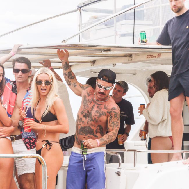 A group of people in swimwear and casual clothes enjoy drinks and conversation on a white boat, with one tattooed man in blue shorts standing near the center. The scene looks lively and relaxed under cloudy skies.