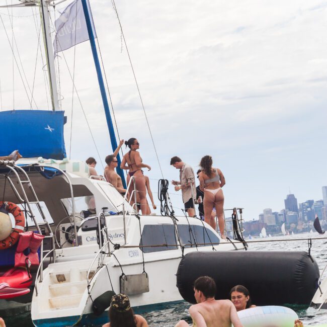A group of people in swimsuits relax on a catamaran and floating mats in the water. The city skyline is visible in the distance under a partly cloudy sky.