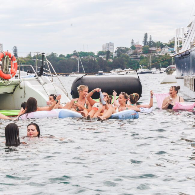 A group of people relax on colorful floaties and swim in the water near two boats, with others socializing on the decks. The scene is lively and festive, set against a backdrop of trees and buildings.
