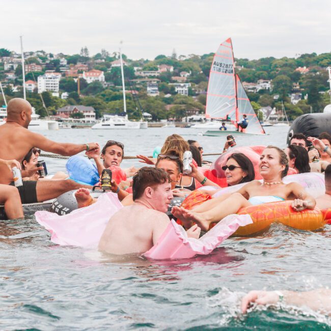 A group of people in swimsuits enjoy a lively pool party in the water with inflatables, drinks, and laughter, with boats and houses visible in the background.