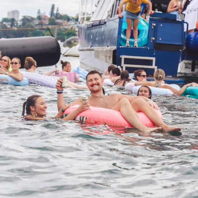People float on inflatable rings in the water near a boat. A smiling man holds cans while relaxing on a pink float, surrounded by others. Some people are about to enter the water from the boat. The scene is lively and playful.