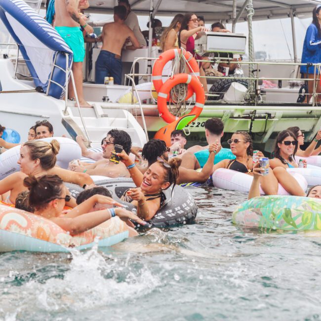 A group of people in colorful floaties relax and socialize in the water near boats during a lively outdoor party. Some hold drinks, and others laugh, enjoying a festive summer atmosphere.