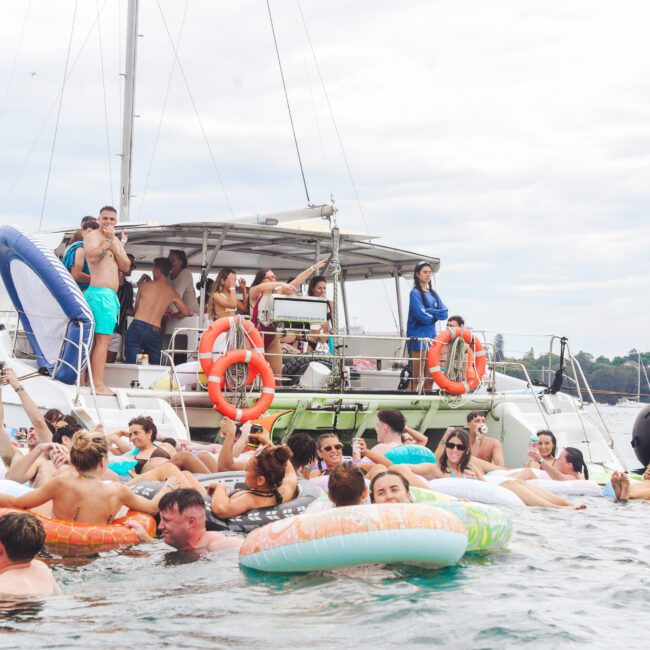 A large group of people in swimsuits relax on colorful inflatable floaties in the water near a boat, enjoying a lively party atmosphere under a cloudy sky.