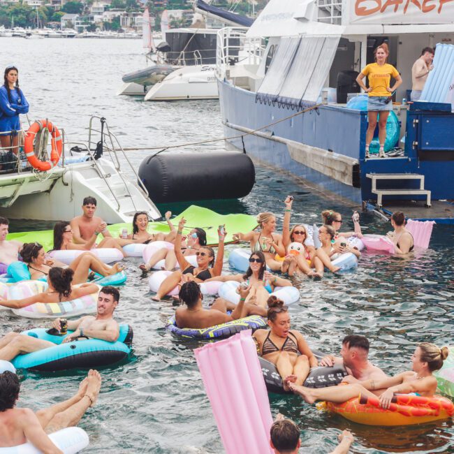 A group of people in swimsuits relax on colorful inflatable floats in the water near boats, enjoying a lively summer party. Some stand on the boats, while others float and chat, creating a festive atmosphere.