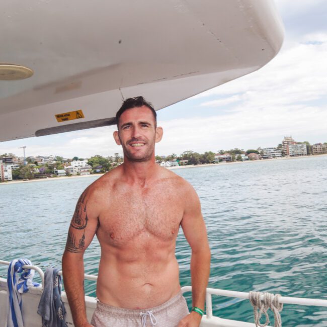 A smiling man stands shirtless on a boat near the railing, with water and a shoreline of buildings and trees visible in the background under a partly cloudy sky.