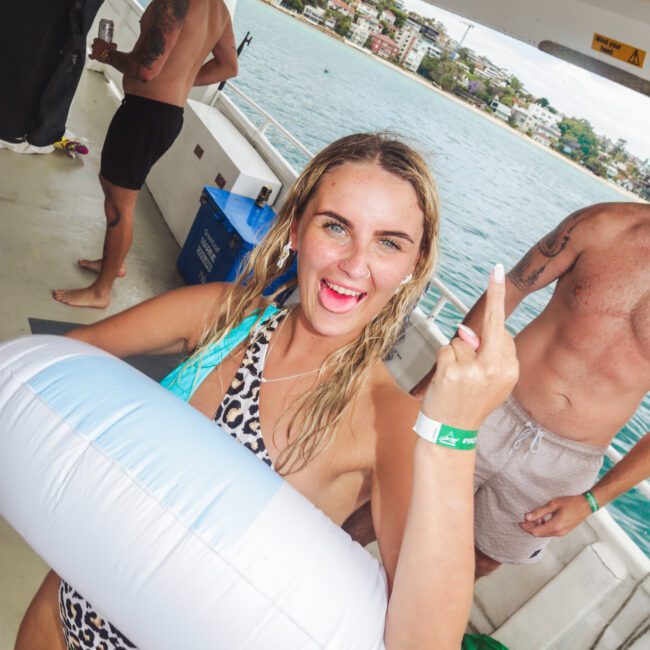 A woman in a leopard print swimsuit and swim ring smiles and gestures with her middle finger on a boat, with other people and seaside houses visible in the background.