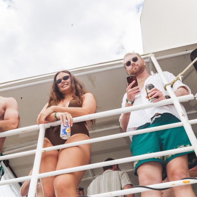 Three people in swimwear stand on a boat deck, holding drinks and looking down over a white railing. The sky is partly cloudy and the atmosphere appears relaxed and casual.