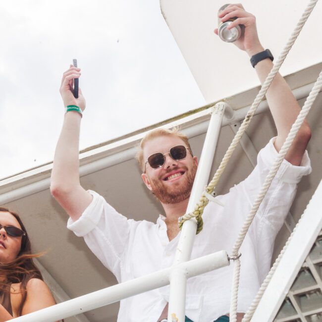 A smiling man in sunglasses raises a phone and a can while standing on a boat deck next to a woman with long hair and sunglasses, who is also holding a can. Both appear to be enjoying themselves.