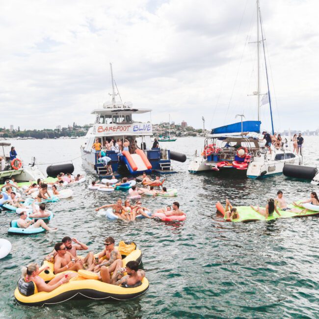 A group of people enjoying a party on boats and colorful floaties in the water, with music, slides, and a lively atmosphere under a cloudy sky near a city shoreline.