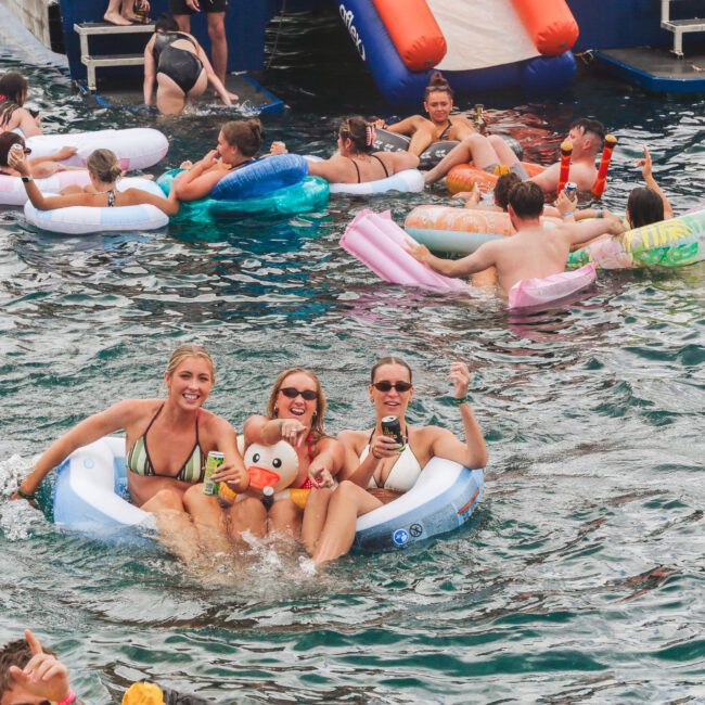 Three women in swimsuits smile and pose on an inflatable float in a crowded pool, surrounded by others on colorful floats. The atmosphere is lively and festive, with people enjoying the water and a floating platform in the background.