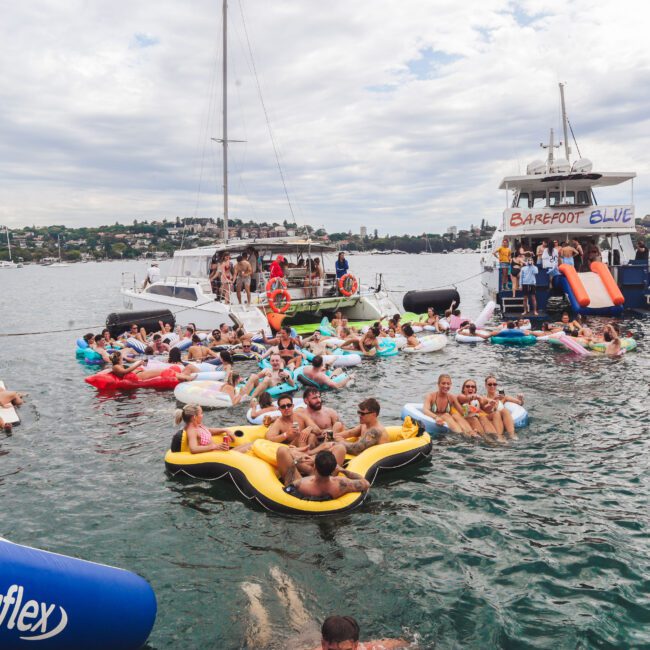 A group of people relaxing on colorful inflatables and swimming near boats in a harbor, with one boat displaying a "Barefoot Blue" sign. The sky is cloudy and land is visible in the background.