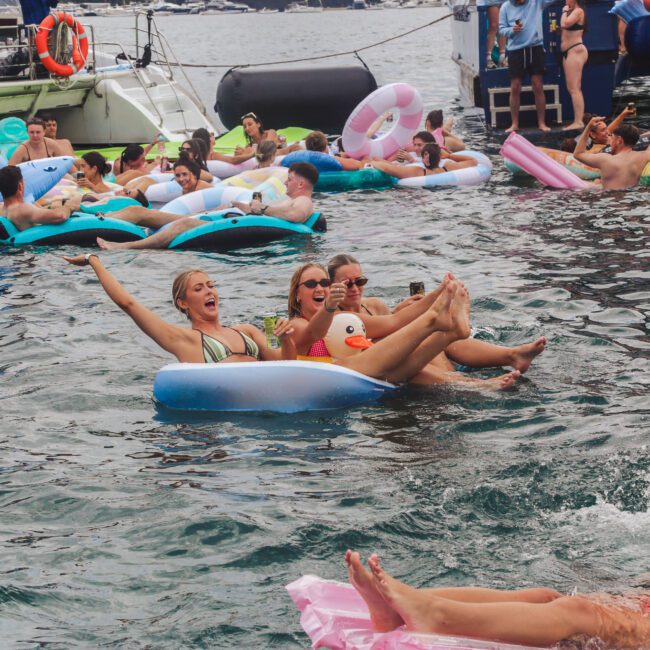 People are relaxing and playing on colorful inflatable floats in the water near boats, smiling and enjoying a lively outdoor pool party.