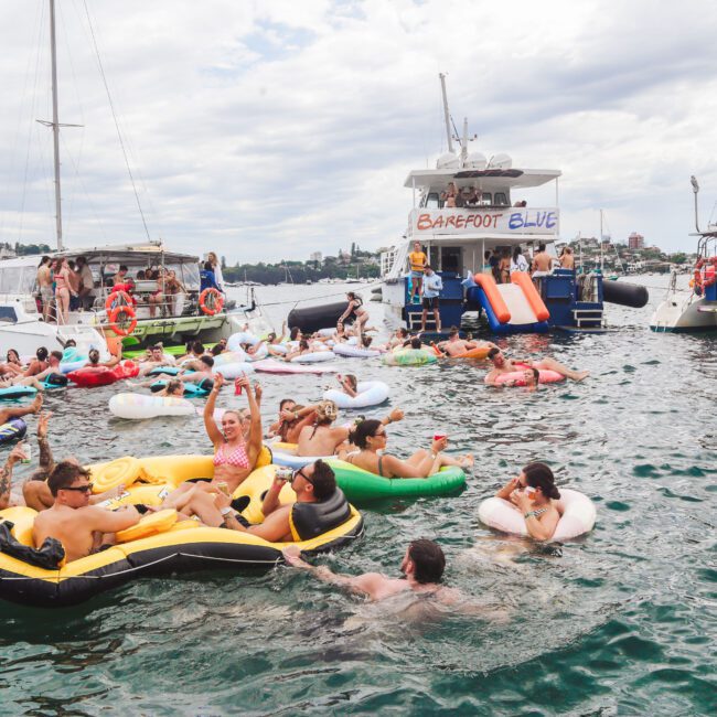 People on inflatable rafts and floats enjoy a lively party in the water near several anchored boats, including one with "Barefoot Blue" on a sign. The scene is festive with many people swimming and socializing.