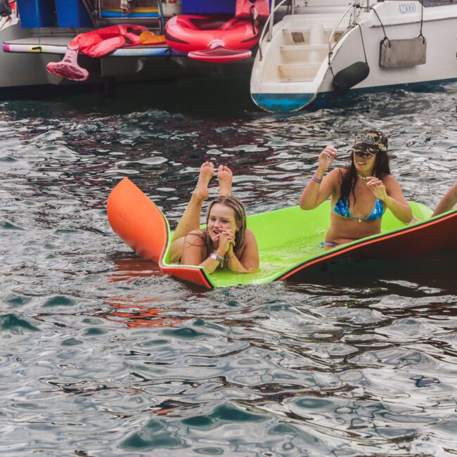 Three young people relax on a floating mat in the water near a catamaran. Two women in swimwear are smiling and one makes a peace sign, while a man sits beside them, all enjoying a day at sea.