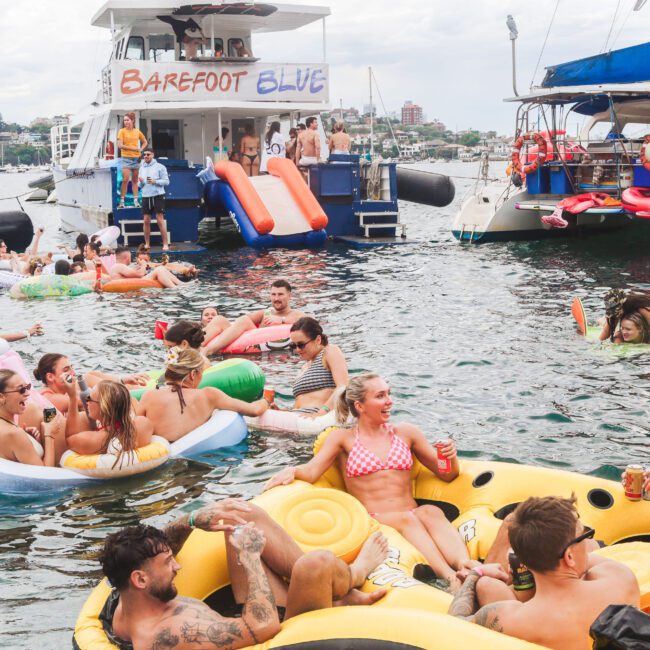 A group of people in swimsuits relax on colorful inflatable floats in the water near two anchored boats, enjoying drinks and socializing at a lively summer party.