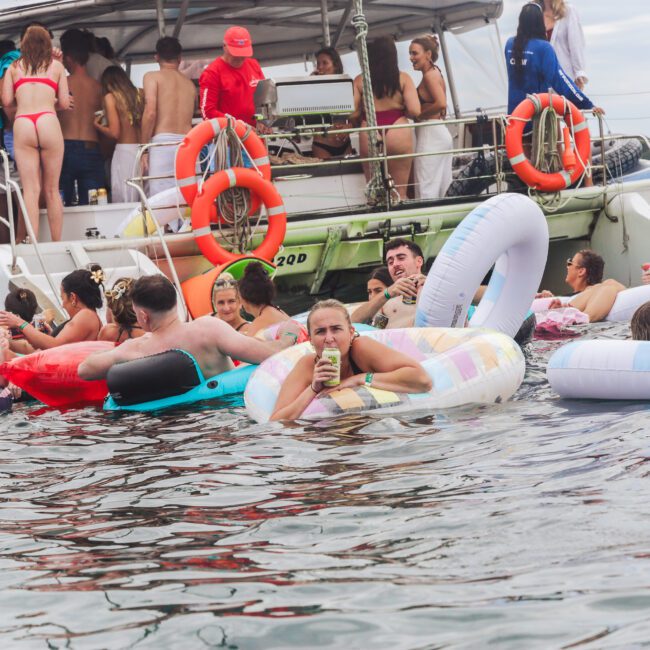 People relax on colorful inflatables in the water near a docked boat filled with more people. Some are talking, swimming, or holding drinks, enjoying a lively, social atmosphere on a cloudy day.