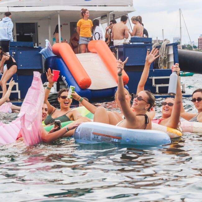 A group of people in swimsuits float on inflatables and celebrate with drinks near a boat, while others stand or sit on the boat’s deck, enjoying a lively summer party on the water.