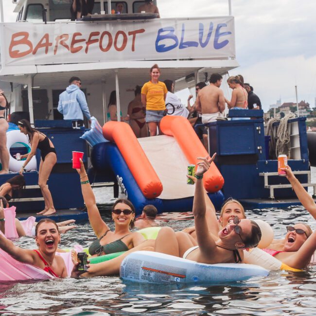 A group of people float on inflatables in the water, smiling and holding drinks, near a boat named "Barefoot Blue" with others onboard. The scene is lively and festive, set near a city shoreline.