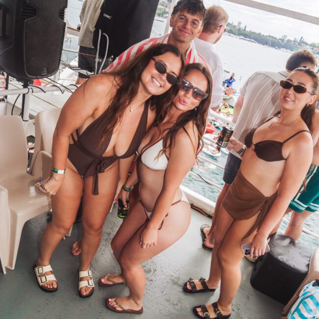 Four people in swimsuits and sunglasses smile and pose on a boat deck at a lively outdoor party near the water, with other people and a city skyline visible in the background.