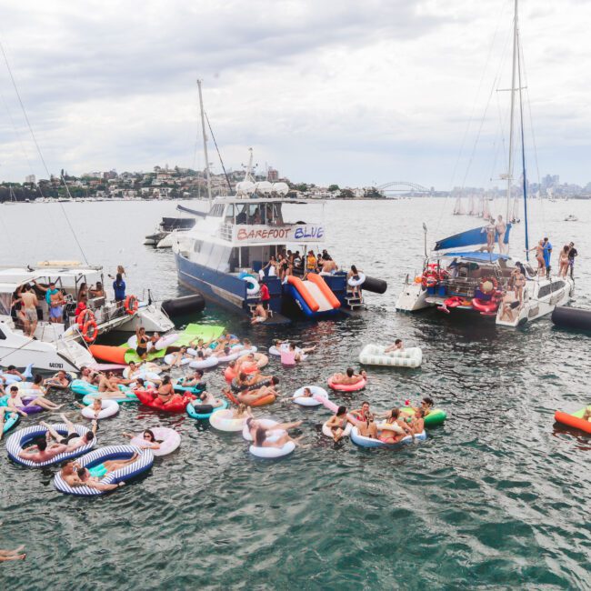 A lively scene of people relaxing on colorful inflatables and swimming near boats gathered close together in a harbor, with a city skyline and cloudy sky in the background.