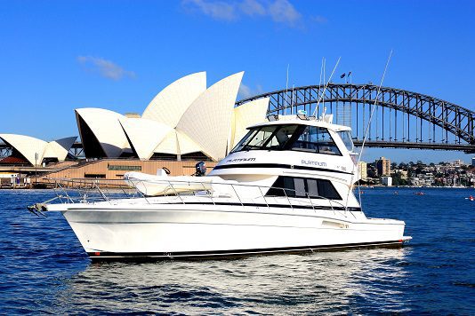 A Platinum yacht glides on the water with the Sydney Opera House and Sydney Harbour Bridge in the background beneath a clear blue sky.
