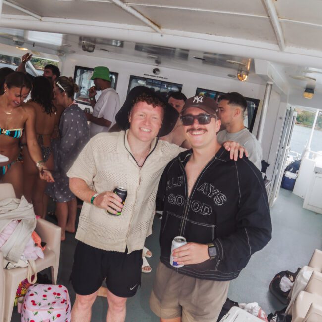 Two smiling men with drinks pose on a boat; one wears a short-sleeve shirt and shorts, the other a black jacket and cap. Other people in swimsuits and beachwear mingle in the background.