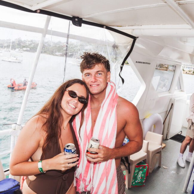 Two people in swimsuits smile and pose with drinks on a boat. The man has a striped towel around his neck, and the woman wears sunglasses. Another person stands in the background, also holding a drink. Water and boats are visible outside.