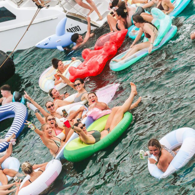 A group of people float on colorful inflatable pool floats in the water near a boat, many smiling, raising drinks, and enjoying a lively party atmosphere.