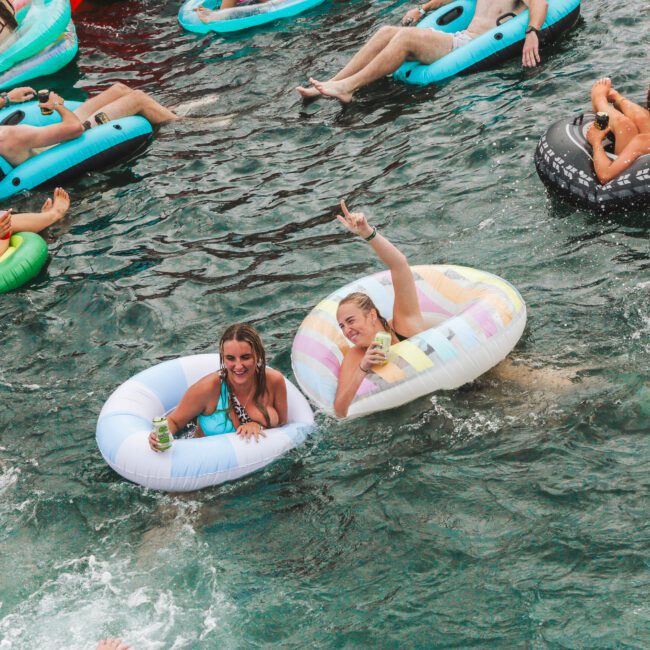 People relaxing on colorful pool floats in a crowded body of water; two women in the center smile and raise drinks while others float nearby, splashing and enjoying the summer atmosphere.