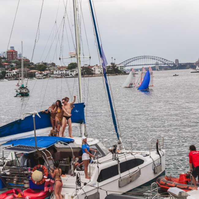 Several people are on a sailboat in a busy harbor, some waving and posing for photos. Other boats and sailboats are on the water, with a city skyline and a large bridge in the background.