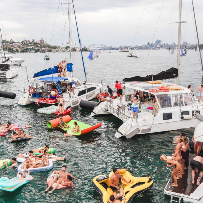 A lively boat party with people on yachts and colorful inflatable floats in the water, set against a city skyline and sailboats under a cloudy sky.