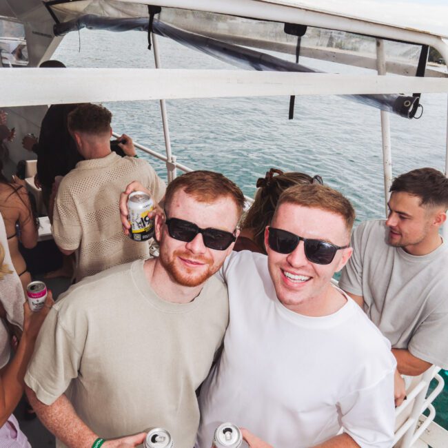 Two men wearing sunglasses and holding drinks smile at the camera on a boat. Other people are in the background, and the boat is on the water under a partly cloudy sky.