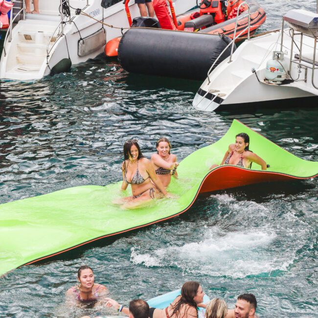 People relax and play on a bright green floating mat in the water near boats. Several individuals swim around them, enjoying a lively and social atmosphere.