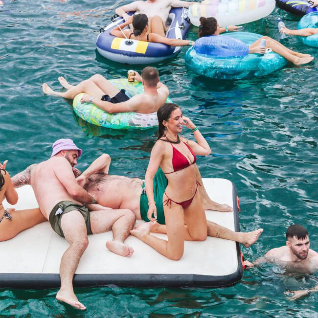 People relax on floats and a large mat in clear blue water. Some are lying down, chatting, or swimming, while others lounge on inflatable tubes, enjoying a sunny day outdoors.