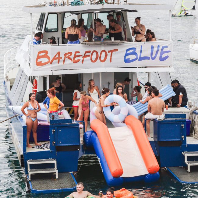 A group of people in swimwear enjoy a party on a boat called "Barefoot Blue," which has an inflatable slide at the back leading into the water. Some are on the slide, others relax or socialize on the boat.