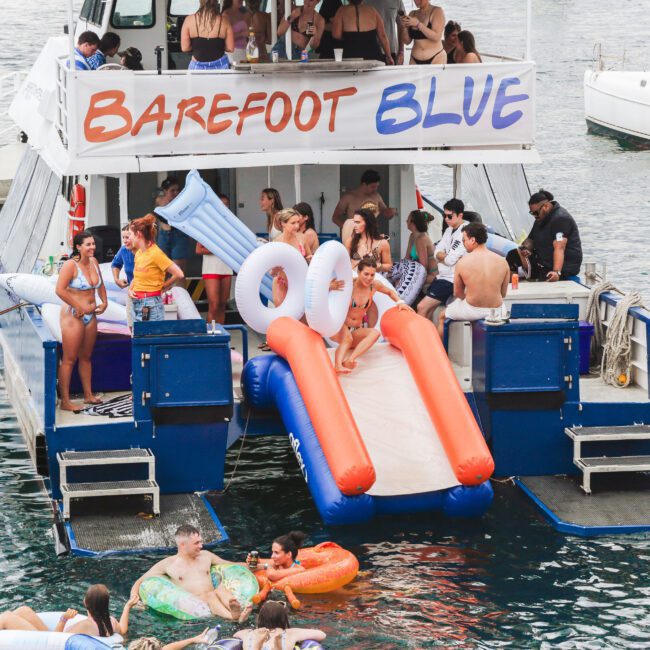 A group of people in swimsuits enjoy a lively party on a boat named “Barefoot Blue,” with some using an inflatable slide into the water, while others relax on floaties in the sea nearby.