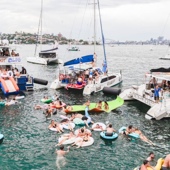 People relax on pool floats and swim in the water near several anchored boats during a lively party on a bay, with city buildings visible in the background under a cloudy sky.