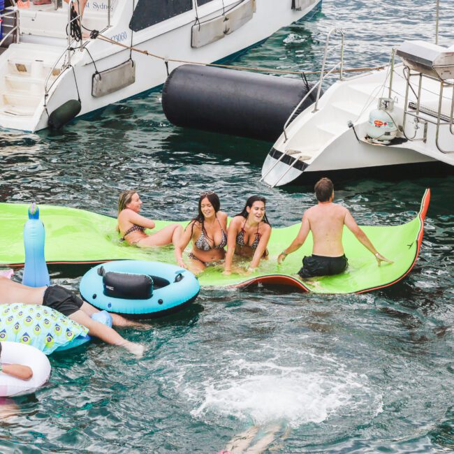 People relax and socialize on colorful inflatables and mats in the water near docked boats, enjoying a sunny day. Some are sitting and laughing, while others float or swim in the lively scene.