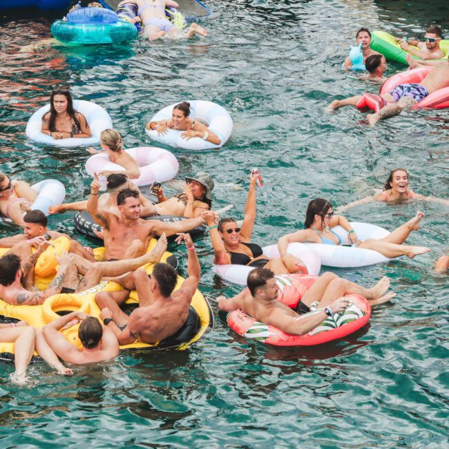 A large group of people relax and have fun on colorful inflatable tubes and floaties in a body of water, enjoying a lively, sunny day together.