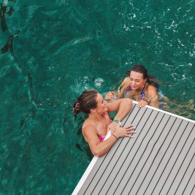 Two women in swimsuits are in clear green water, smiling and holding onto the edge of a wooden dock or platform, enjoying a swim together.